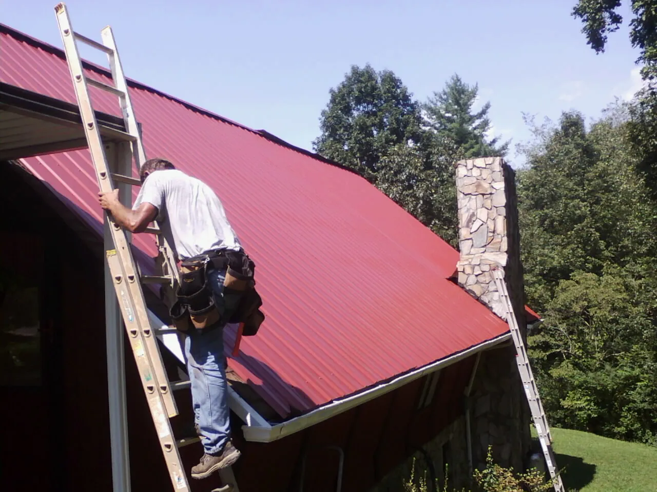Roofer on a ladder beside a red metal roof and stone chimney — Soffit & Fascia Repair by Roofing Brothers in Stephentown